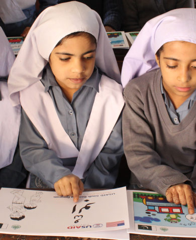 Image of three girls sitting at a school desk and pointing at words on individual learning materials marked "USAID."