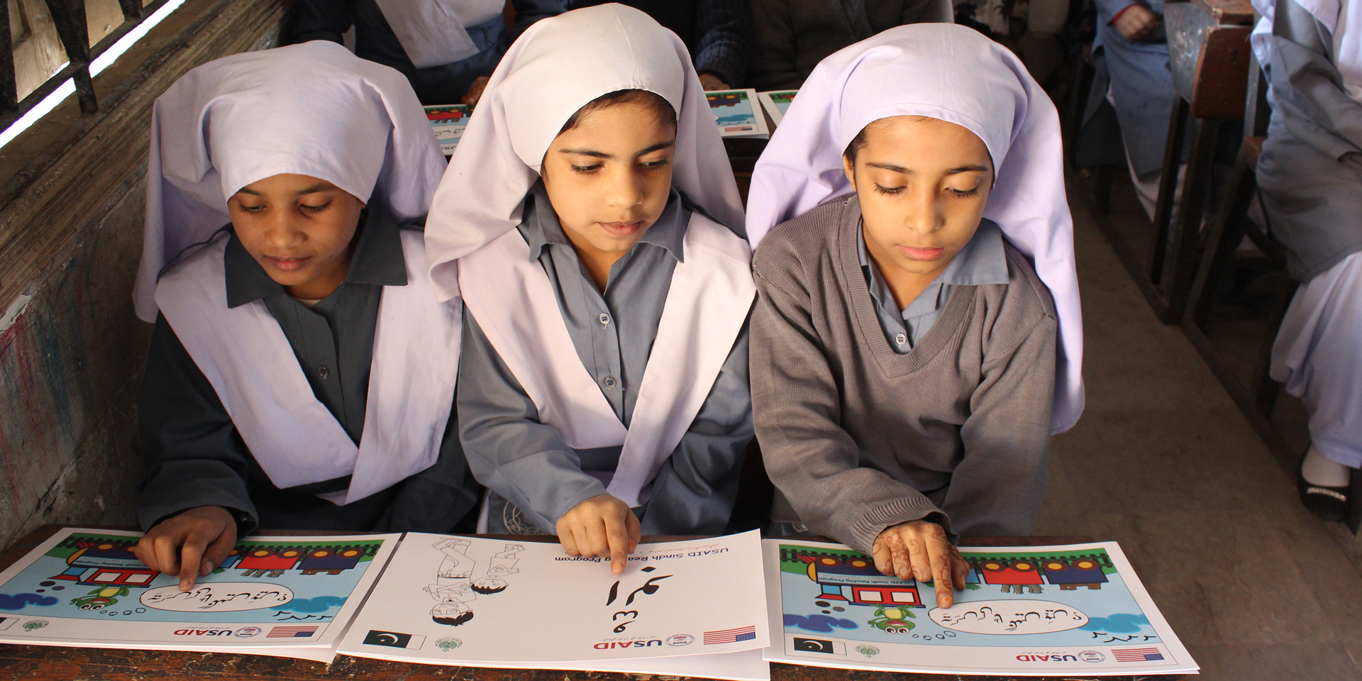 Image of three girls sitting at a school desk and pointing at words on individual learning materials marked 