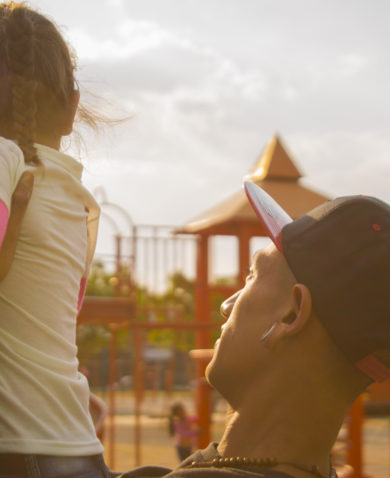 Image of a man in a baseball cap holding up a small girl in a playground.