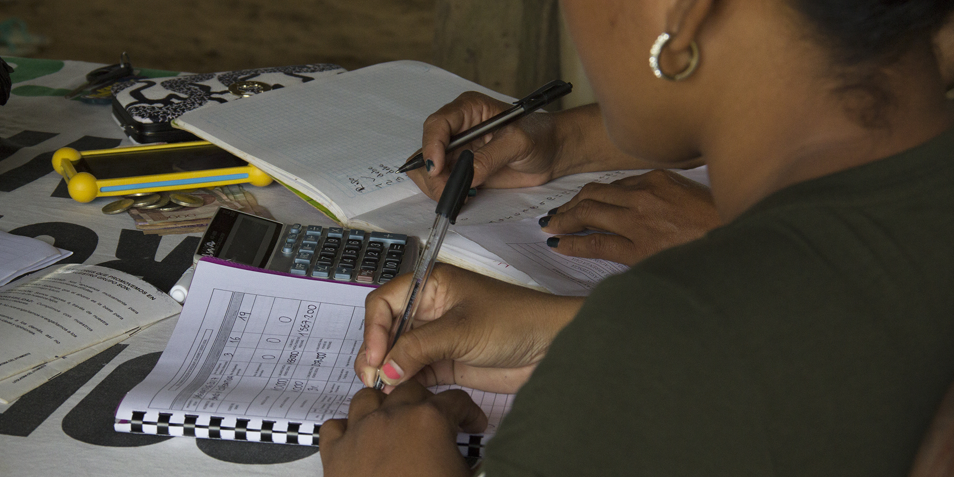 Image of two people sitting at a table and writing into workbooks with a calculator between them.