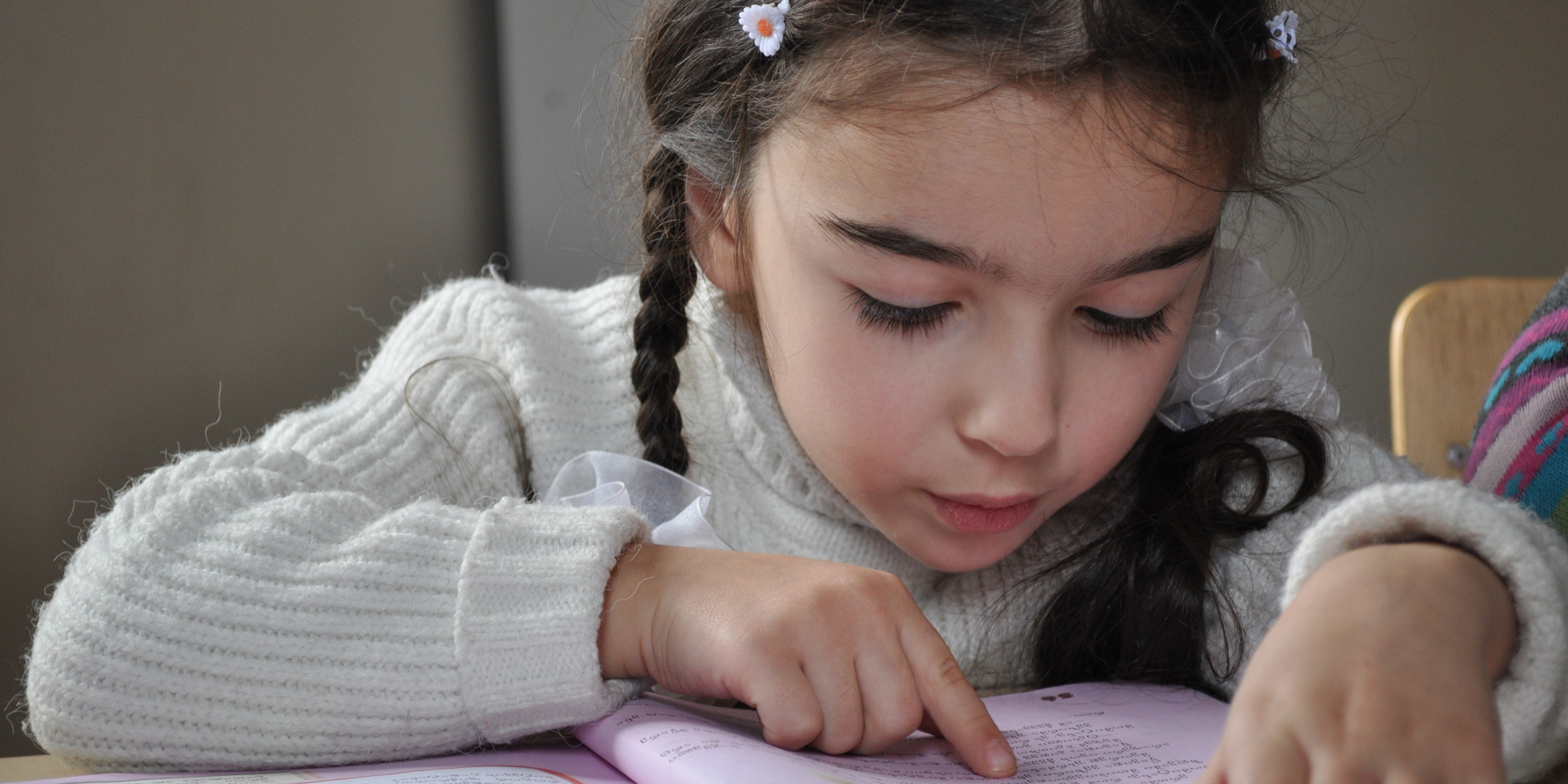 A young girl sitting at a school desk and reading from a purple-colored book.