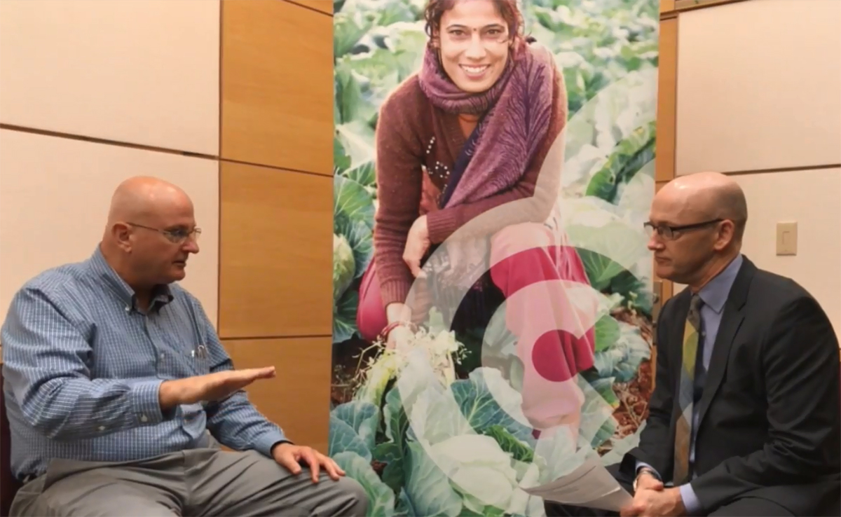 Image of two men sitting on either side of a large banner and speaking. The banner shows a smiling woman kneeling amidst several growing cabbages.