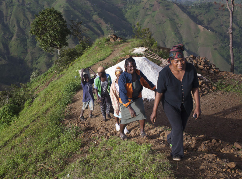 Five members of the Furcy Association of Women Farmers walking up a thin road on a hill. Several mountains can be seen in the distance.