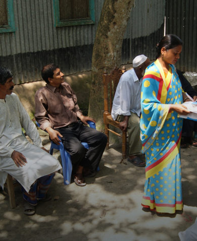 Outside, a group of people sits in chairs in a circle as a woman in the middle hands out flyers. A man at the far end is giving a presentation.