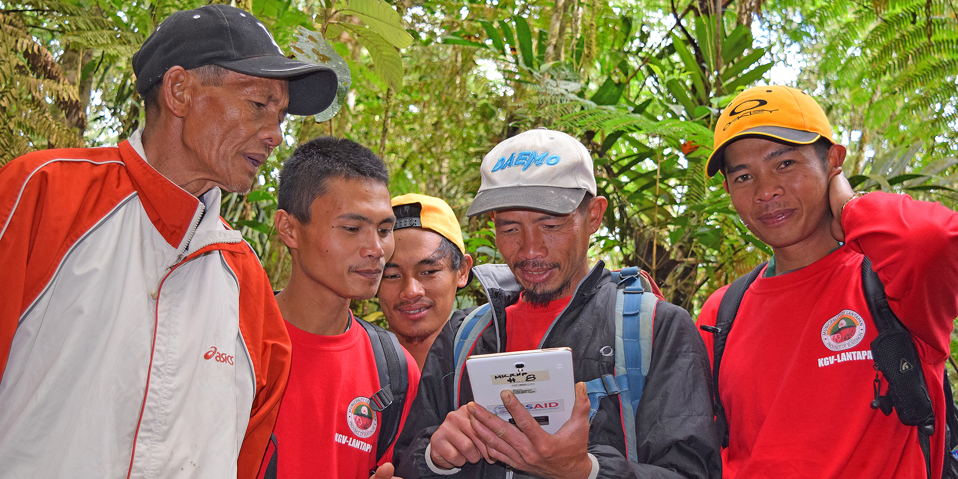 A man showing a tablet he is holding to four other men standing alongside him. They are in a green forest.