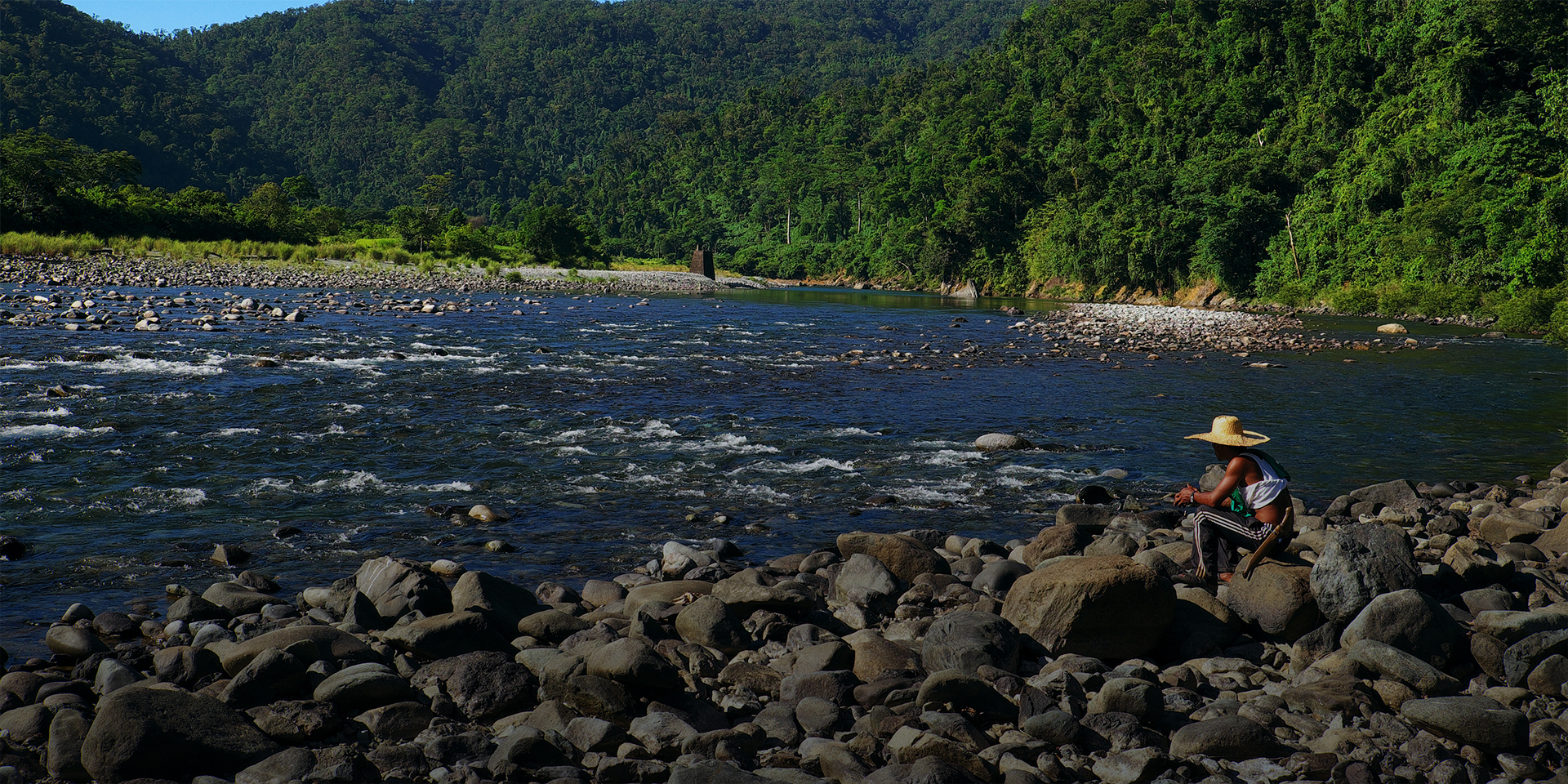 A person sitting on an outcrop of rocks in front of a flowing river. A mountainous landscape covered with trees is in the distance.