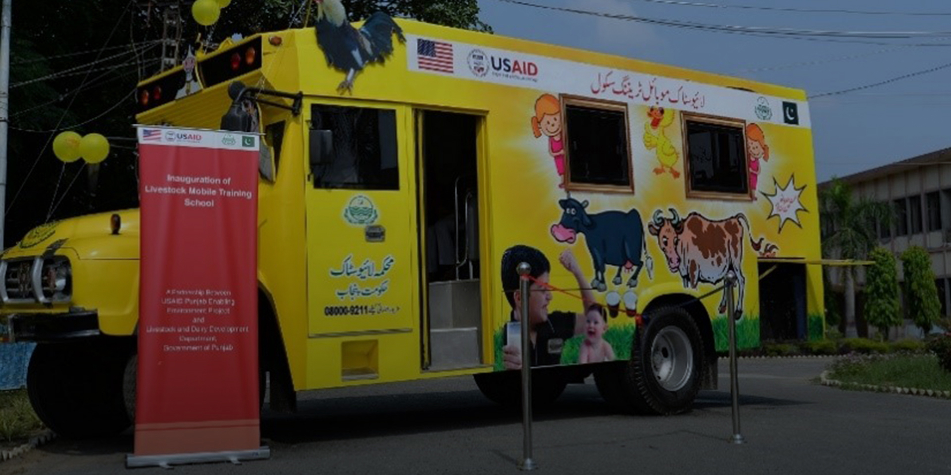 Image of a truck in the shape of a small school bus with cartoon images of cows and children on the side. Beside the truck is a sign that reads 