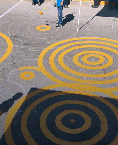Image of two people painting the outlines of concentric circles with orange paint using a long-handled paint roller.