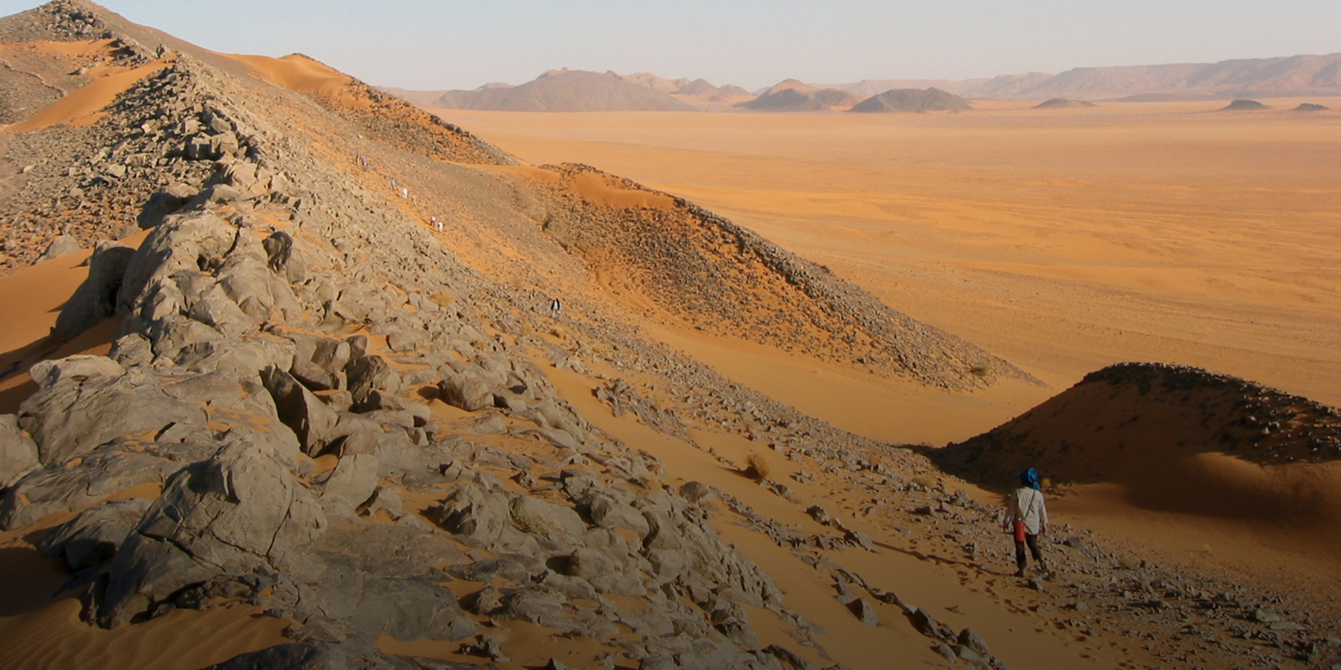 A mountainous desert with hills in the distance. A line of people can be seen walking along the base of a hill.