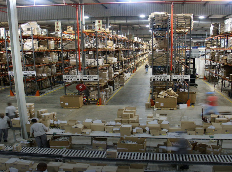 Image of a large warehouse with workers packing cardboard boxes in the foreground and several aisles of steel shelves holding boxes in the background.