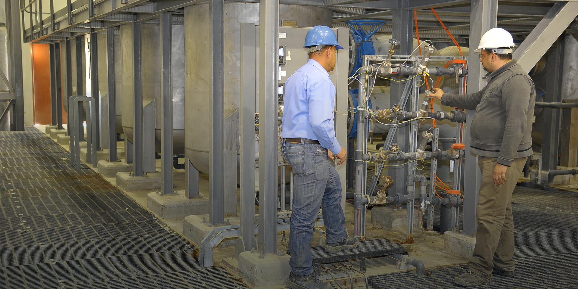 Two men in hard hats inspecting several pipes and valves in a water treatment facility.