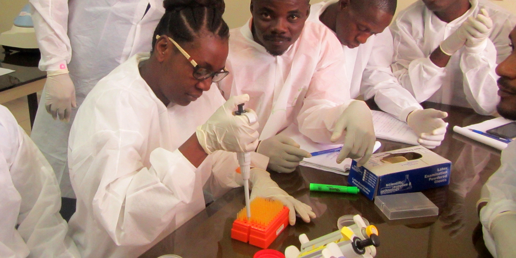 Image of several lab technicians sitting at a large desk as one injects fluid into a test tube.