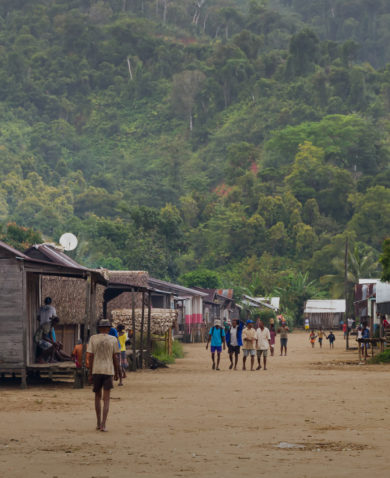The dirt road of a village with several wooden structures on each side. People are walking up and down the street. A large hill covered with trees is in the background.