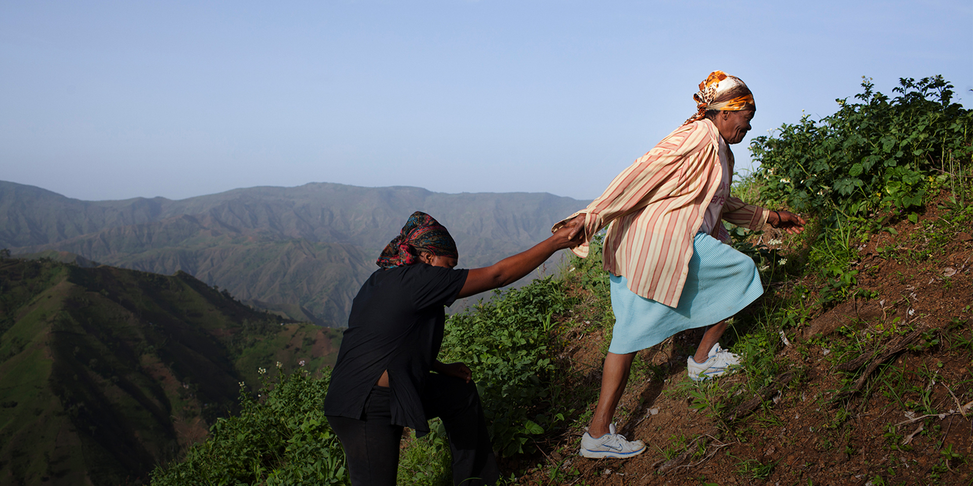 Two women walking up a steep hill. The one in front is helping the other behind her by providing her hand as leverage.