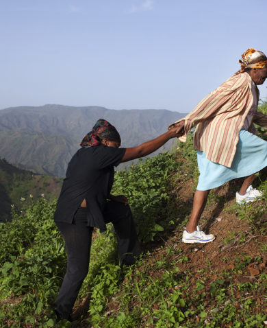 Two women walking up a steep hill. The one in front is helping the other behind her by providing her hand as leverage.