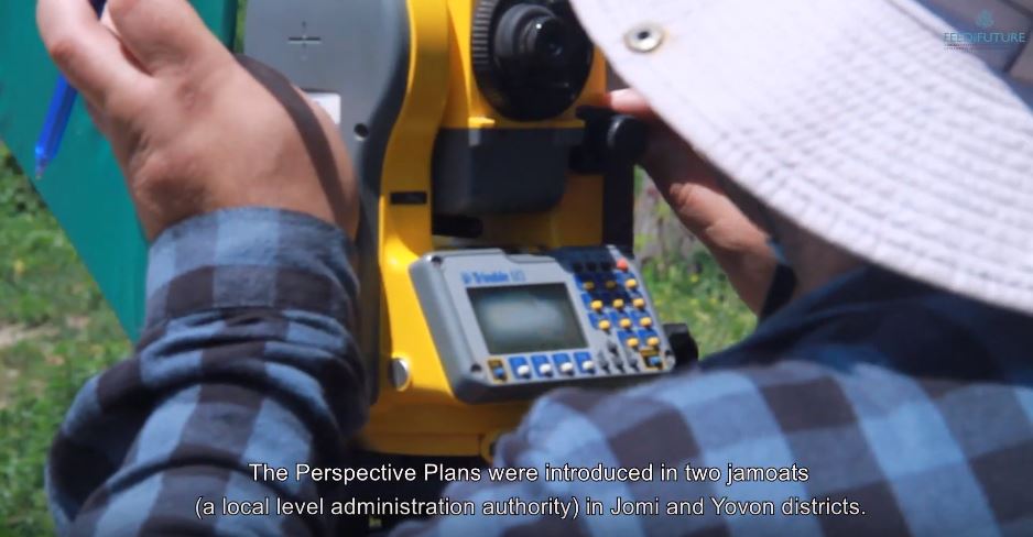 A closeup image of a survey device being operated by someone in a field. Below is a closed caption that reads "The Perspective Plans were introduced in two jamoats (a local level administrative authority" in Jomi and Yovon districts."
