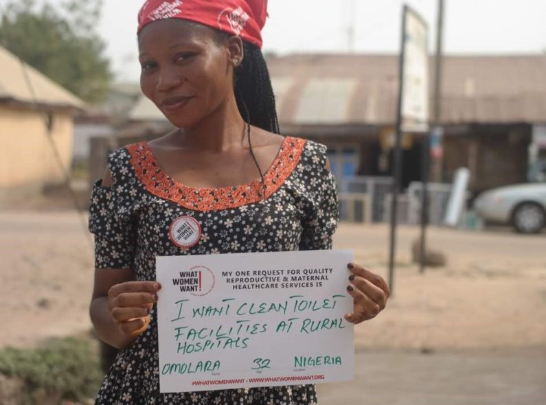 Image of a woman standing in a village. She is smiling and holding a sign that says "I want clean toilet facilities at rural hospitals."