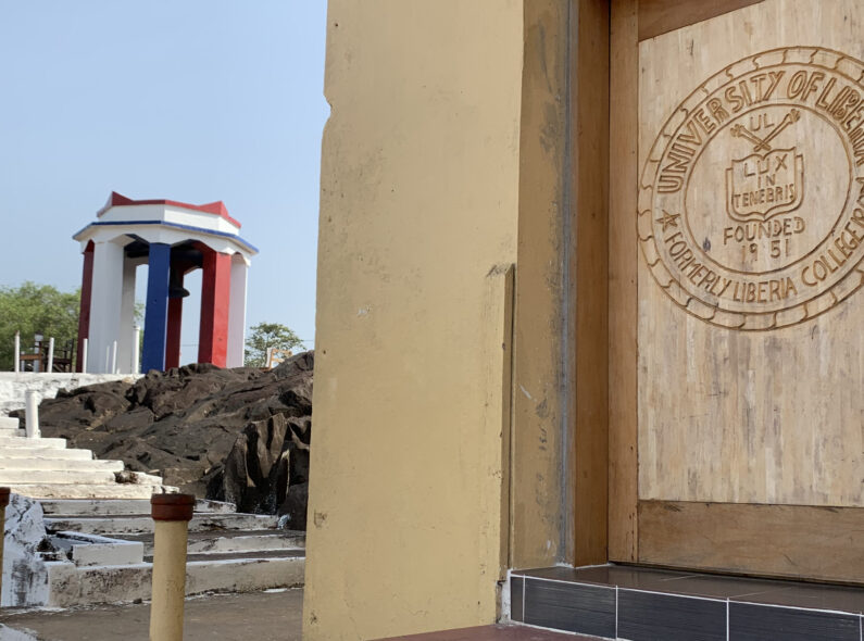 Image of the logo for the University of Liberia carved into wood on a door. Beside it is a stairway leading up to a gazebo.