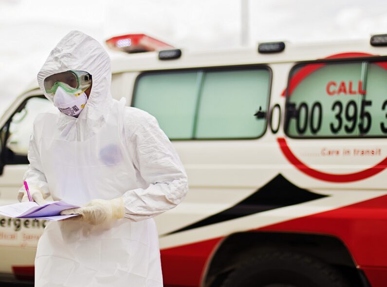 A person in a hazmat suit standing beside an emergency vehicle and holding a set of documents.
