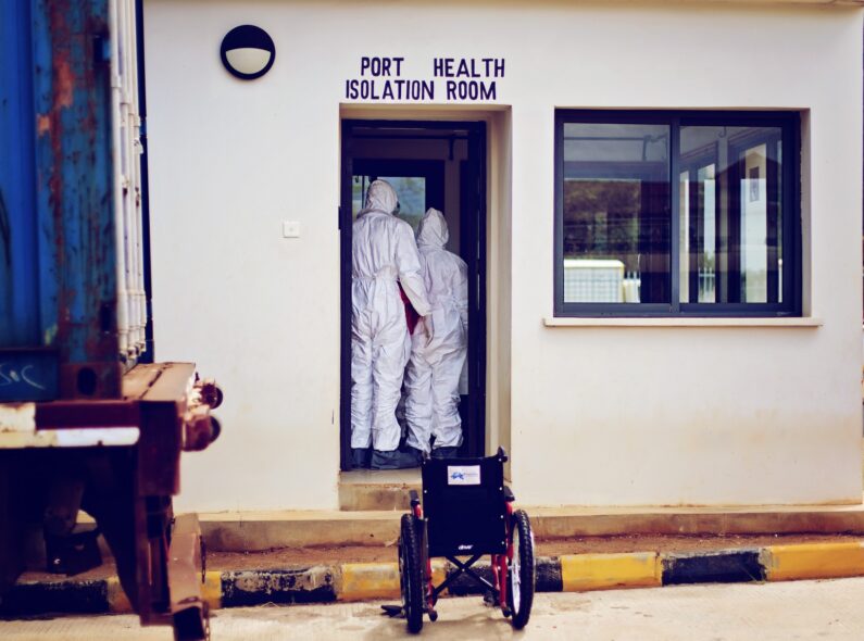 Image of two people in hazmat suits standing inside a doorway with "Port Health Isolation Room" written above.