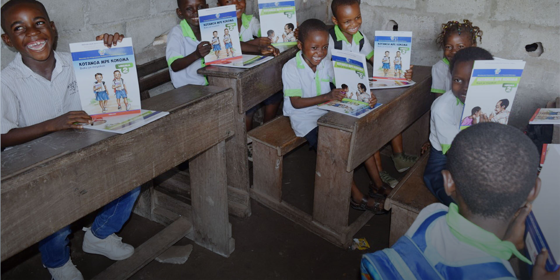Image of children sitting at school desks and smiling as they hold up activity books.