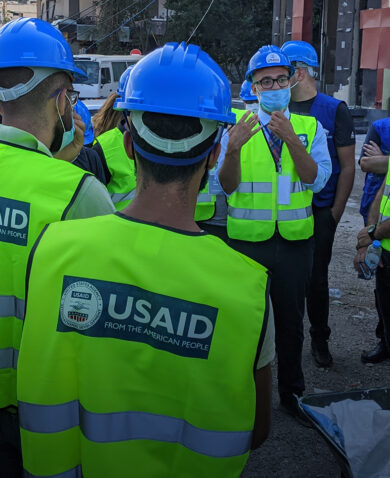 Several workers wearing hard hats and reflective vests marked "USAID" stand and listen to a foreman speaking.