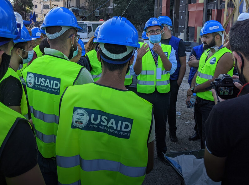 Several workers wearing hard hats and reflective vests marked "USAID" stand and listen to a foreman speaking.