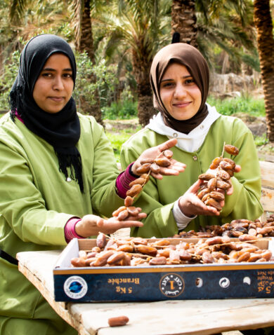 Women smiling and sitting at a table outside as they sort dried dates.