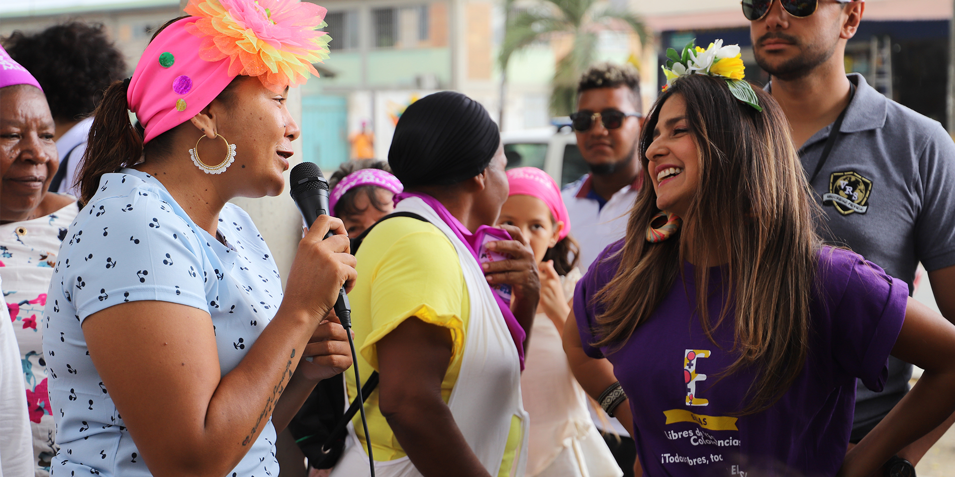 A group of several people gathered outside as a woman with a microphone interviews a smiling woman standing beside her.