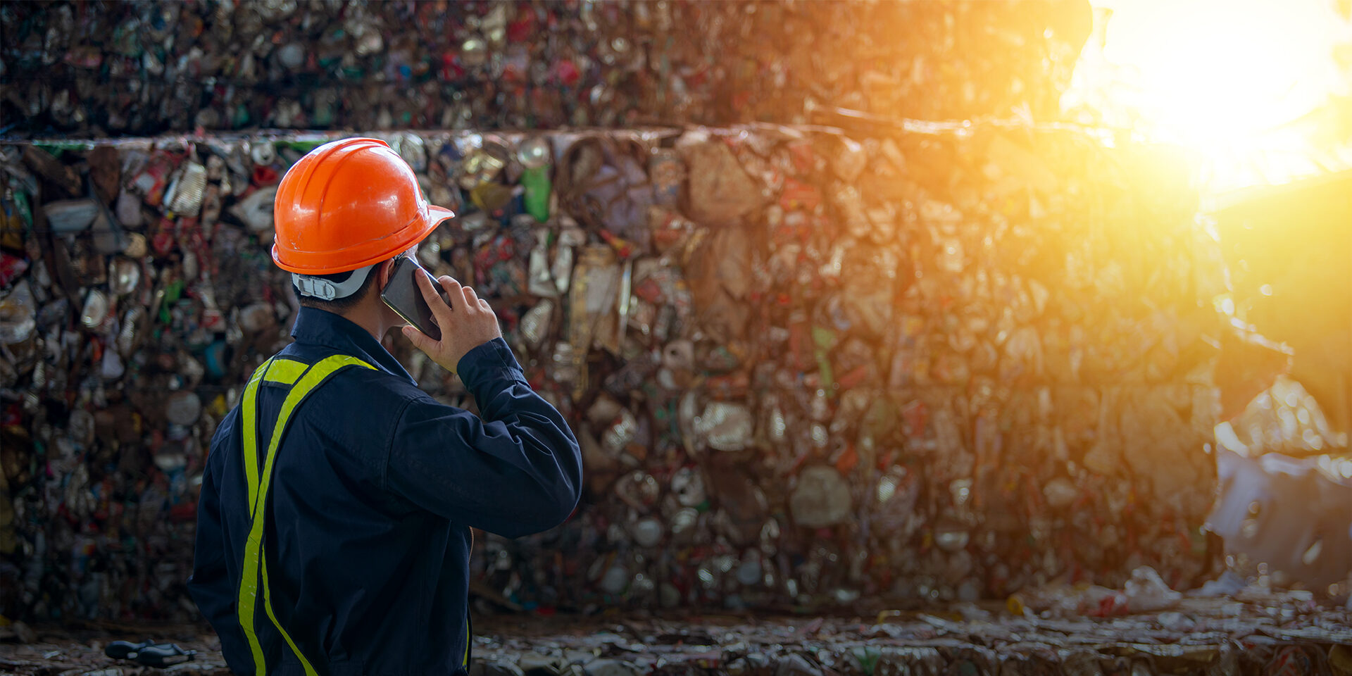A man in a hard hat standing next to a large wall of recycled materials and speaking on a cell phone.