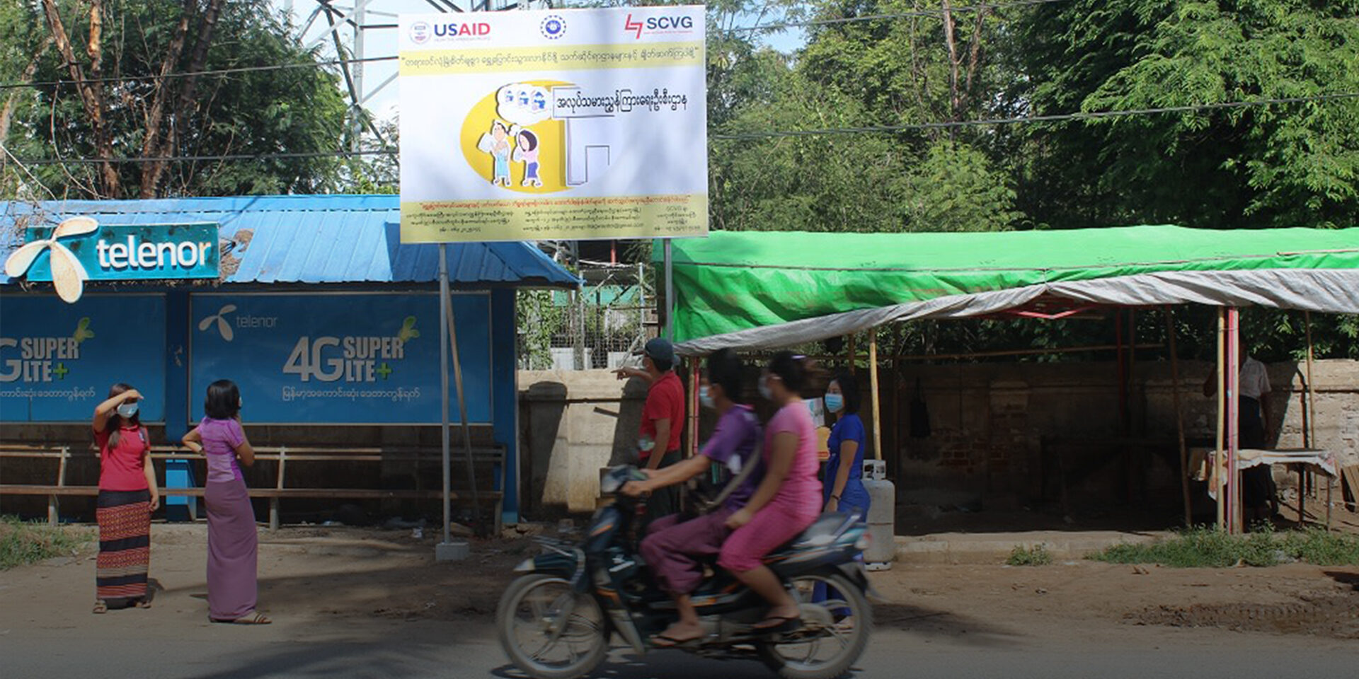 Passengers on a motorbike pass a USAID PRLM billboard about preventing and reporting human trafficking.