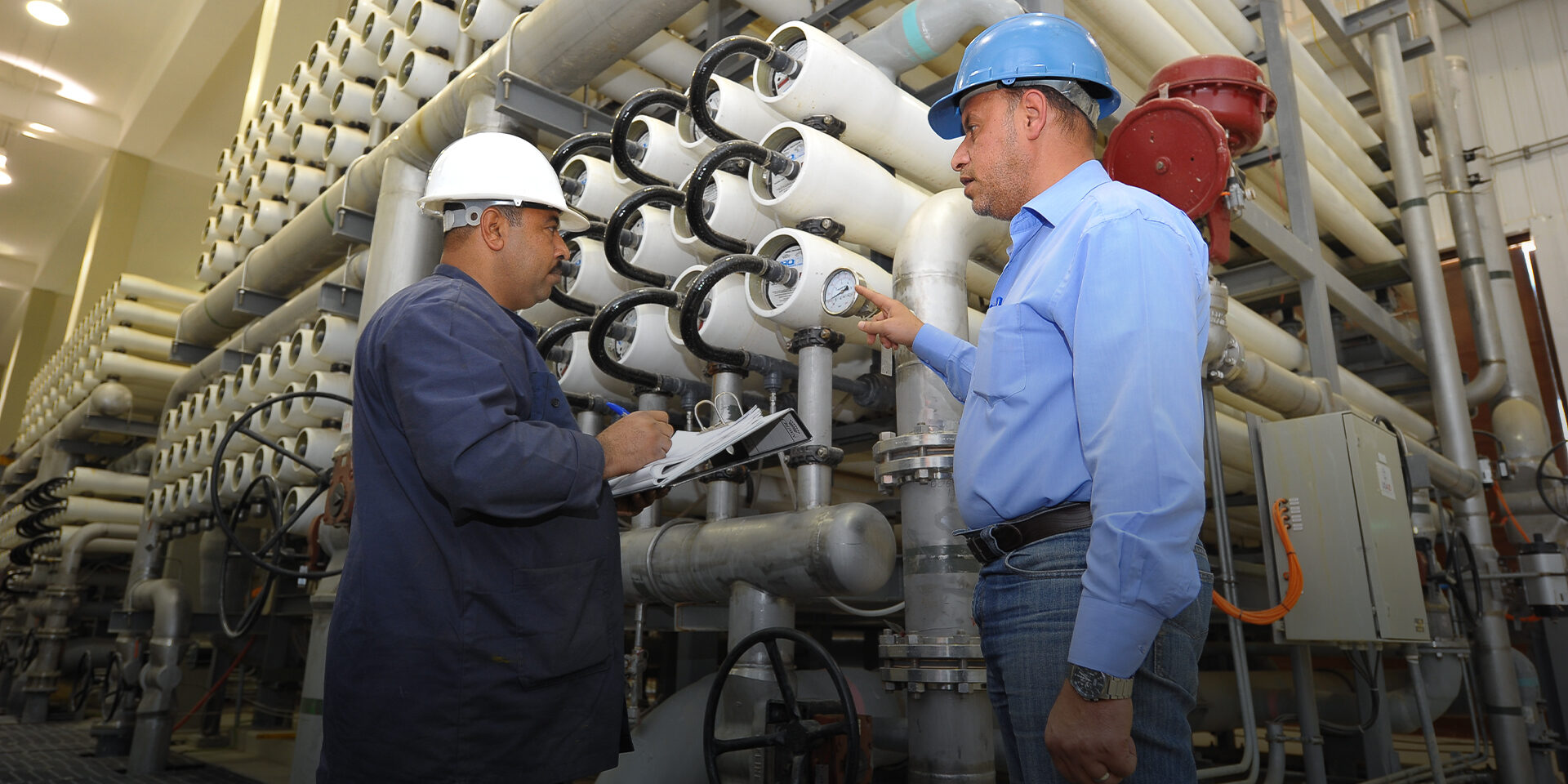 Two men wearing hard hats and standing next to a large machine comprised of pipes and tanks. One is pointing out a reading on a meter.