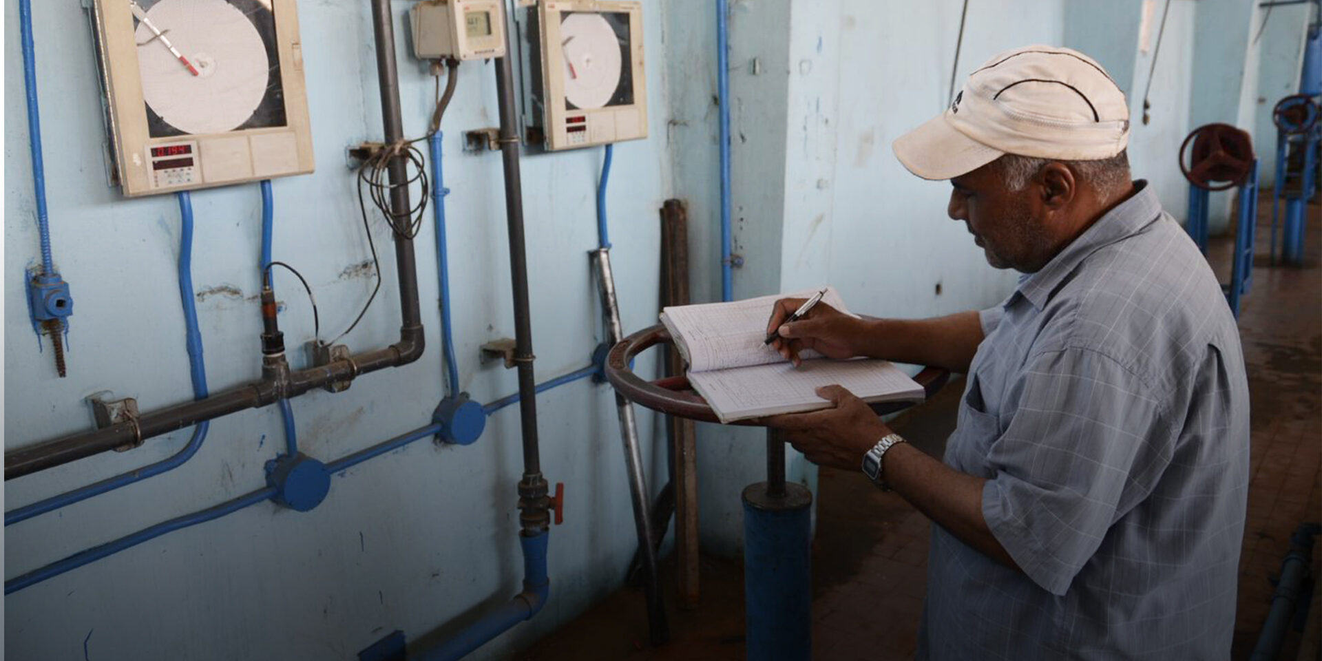 A man writing into a ledger placed on a large valve in a pump station.