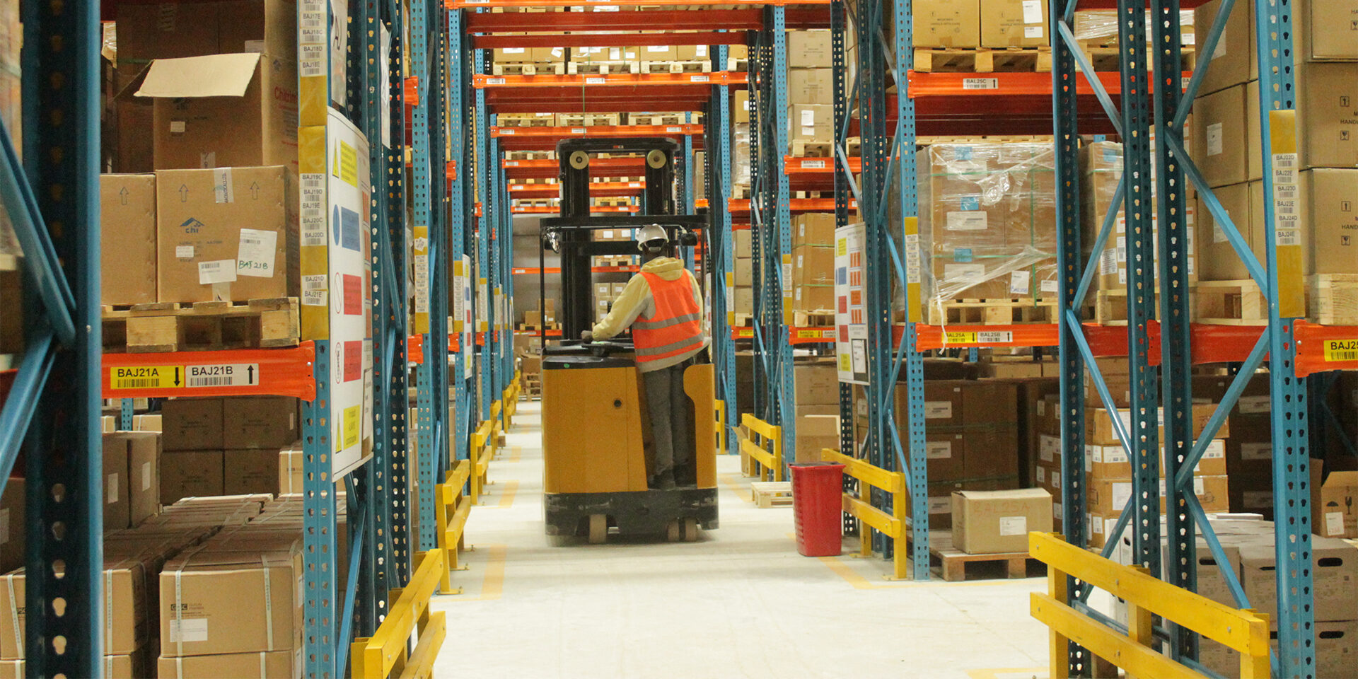 A man driving a forklift down a warehouse aisle with shelves of boxes on each side.
