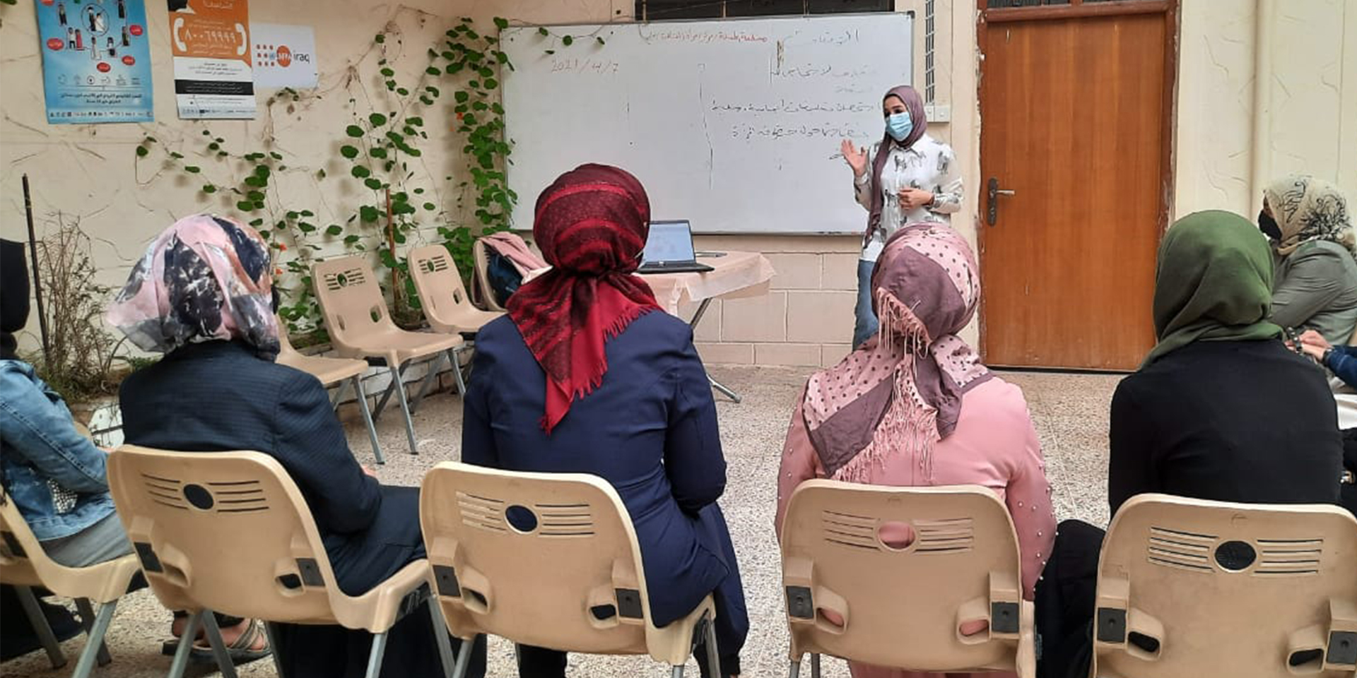 Image of several women in a classroom sitting in chairs and listening to a presentation led by a woman standing next to a whiteboard.