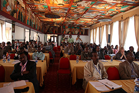 Several people in a conference hall sitting at tables and listening to a presentation.