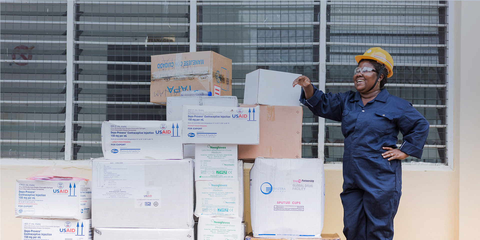 A woman in a hard hat smiling as she stands next to a stack of boxes marked "USAID."