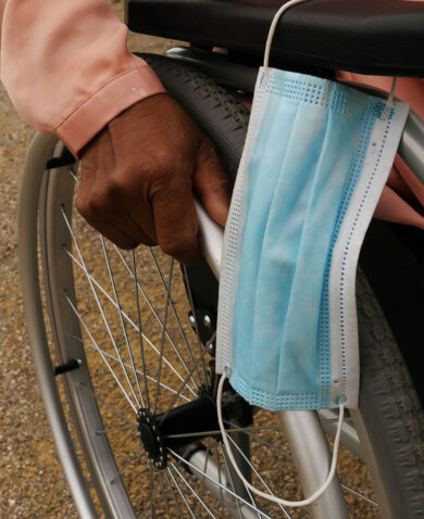A close-up image of a person in a wheelchair with a surgical mask hanging from the armrest.