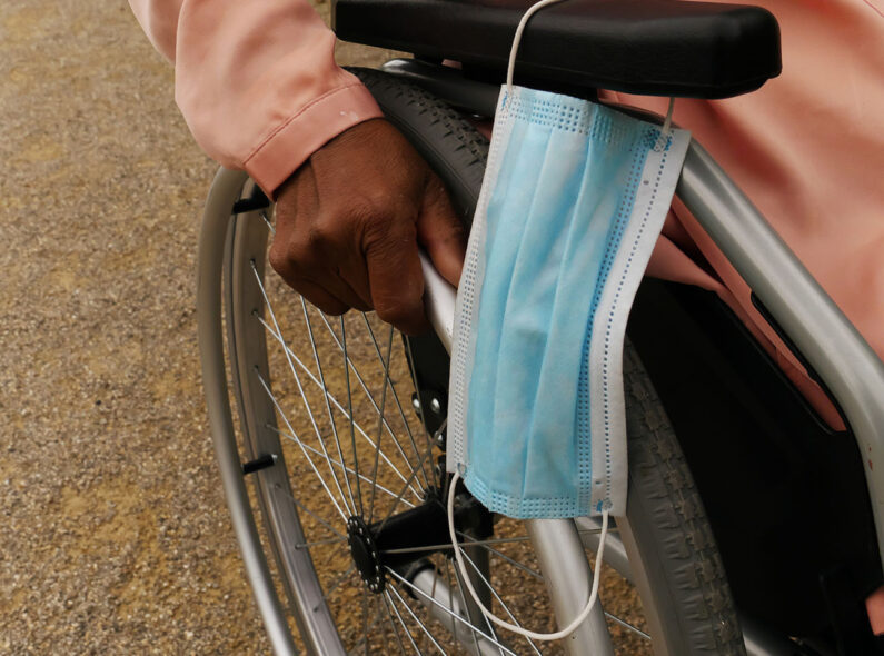 A close-up image of a person in a wheelchair with a surgical mask hanging from the armrest.