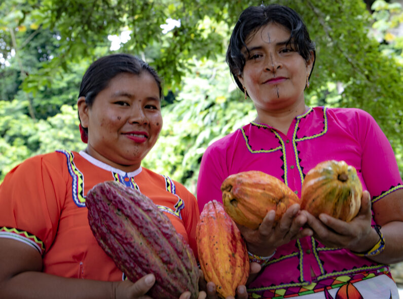 A pair of women posing for a photo outside and holding large orange and purple fruits.