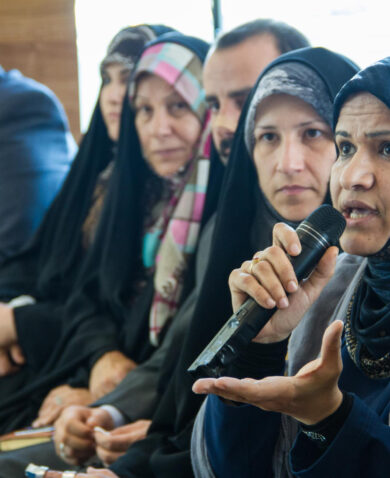 An Iraqi woman wearing a headscarf speaks into a microphone at a meeting.