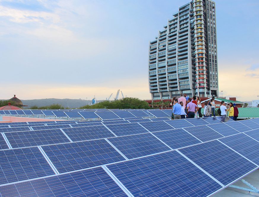 A large group of people standing in the middle of a solar array. A large skyscraper can be seen in the distance.