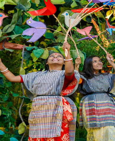 Two young women holding paper birds are part of a performance portraying life inside a forest, in front of the audience attending the launch event of the USAID Ecosystems/Protibesh Activity.