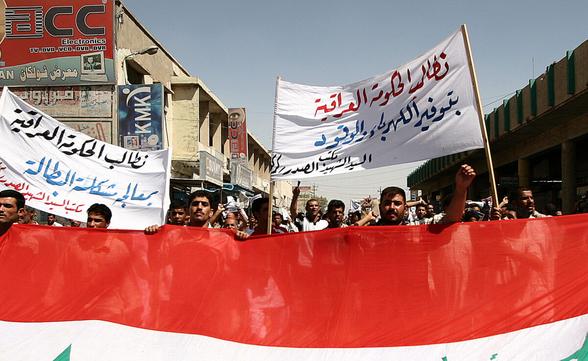 Several people marching in a street holding a large Iraqi flag and several banners.
