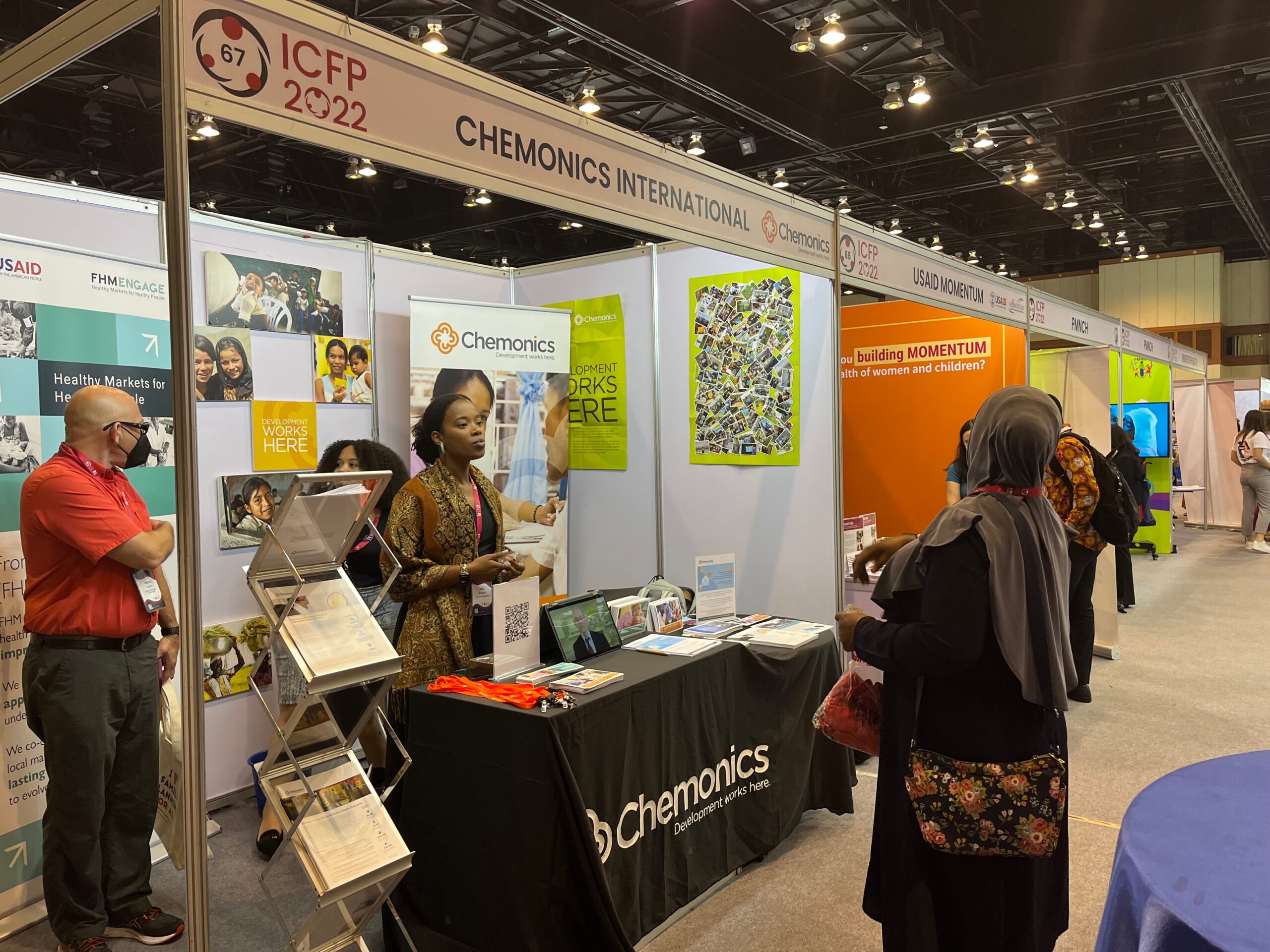 Image of a booth for Chemonics International at a convention. A woman tending the booth is speaking to a visitor.