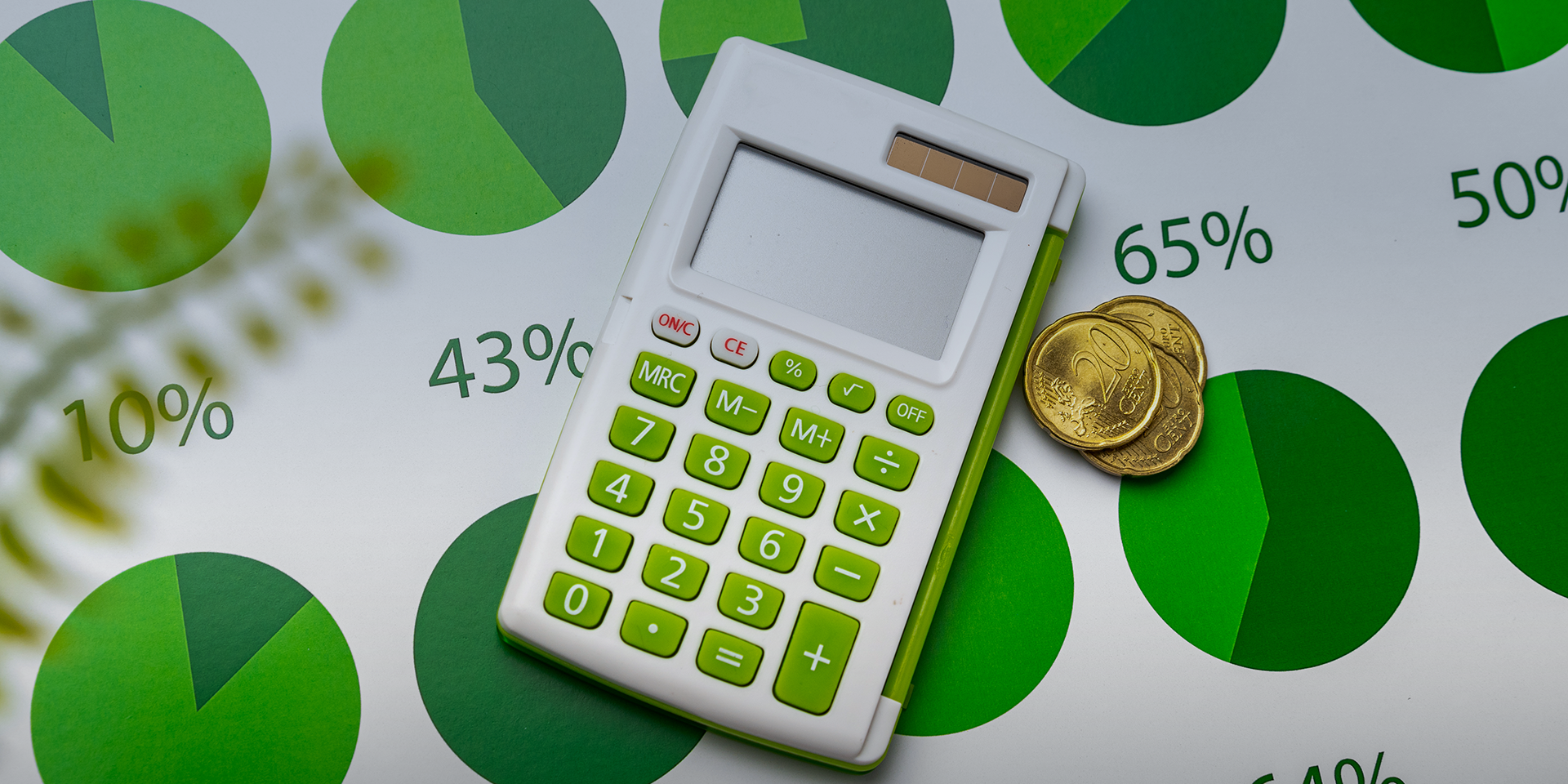 A calculator sits on a table on top of green-colored pie-charts and coins.