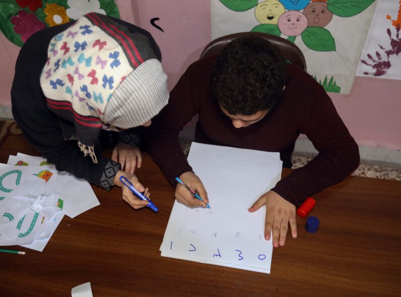 A teacher leaning beside a student at a table who is writing out the alphabet.