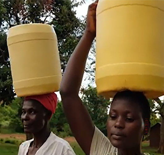 Two women are walking, carrying yellow jugs of water on their heads.