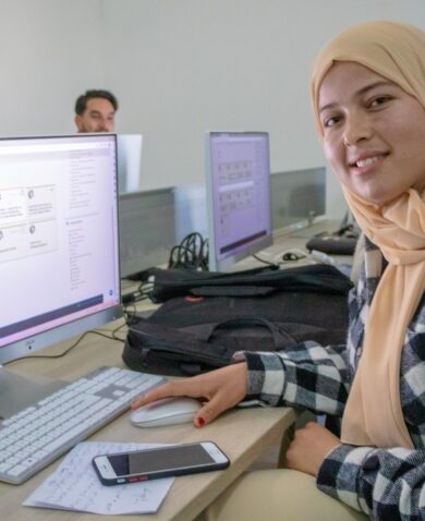 A young woman wearing a head scarf sits in front of a computer and turns to face the camera