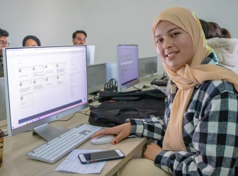 A young woman wearing a head scarf sits in front of a computer and turns to face the camera